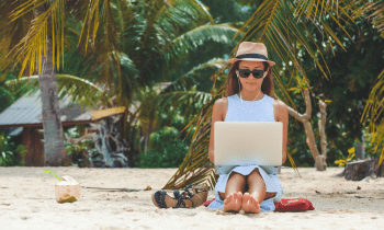 woman working on the beach with her laptop wearing a hat and sunglasses