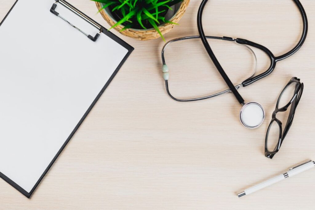 A remote healthcare worker's home office desk, with a stethoscope, clipboard, and plant.