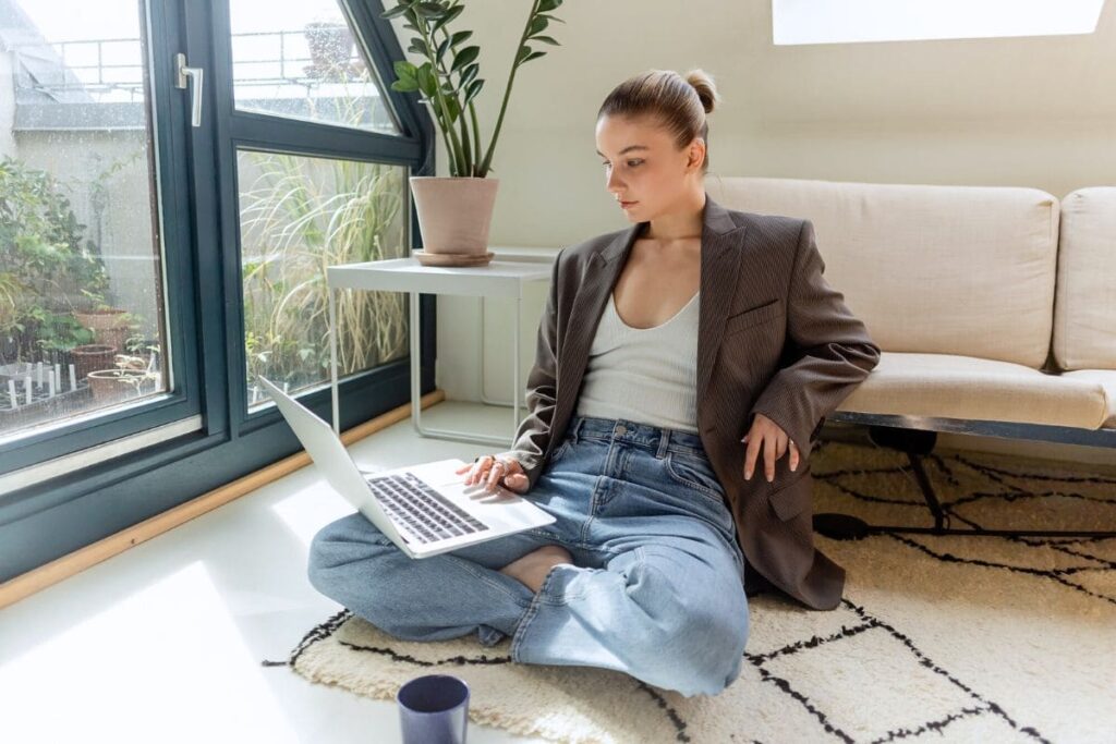 A woman sitting on the floor in her living room and working from home on a laptop.