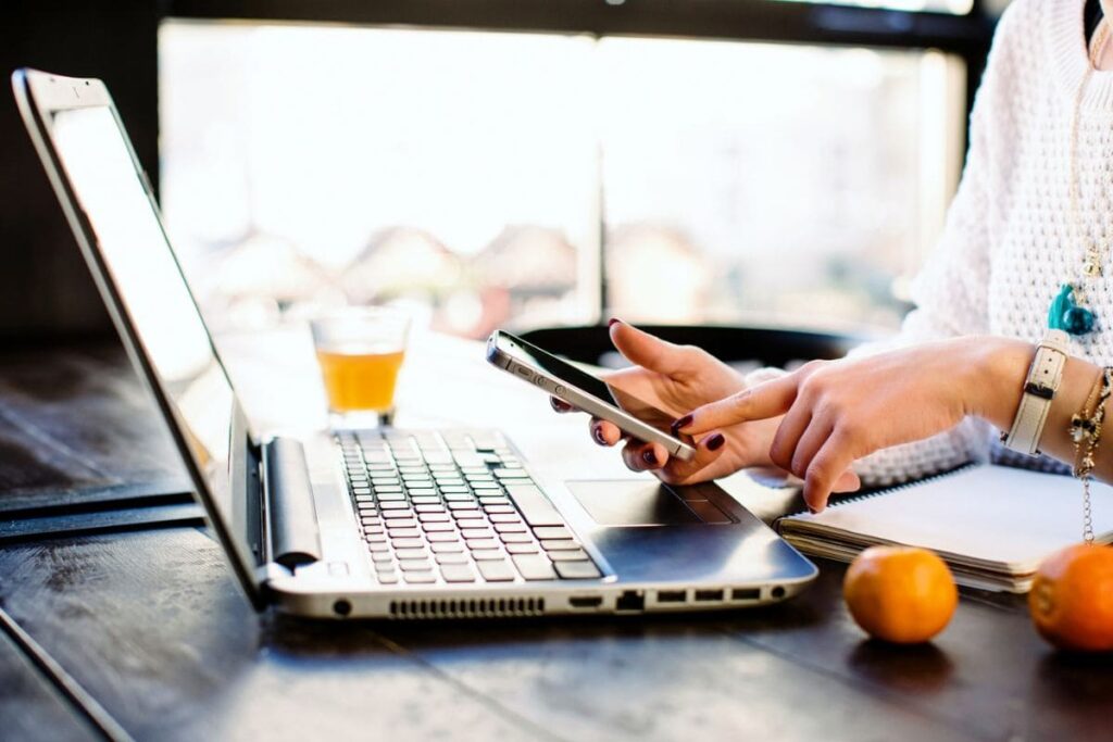 A woman holding a smart phone while working on a laptop at home.