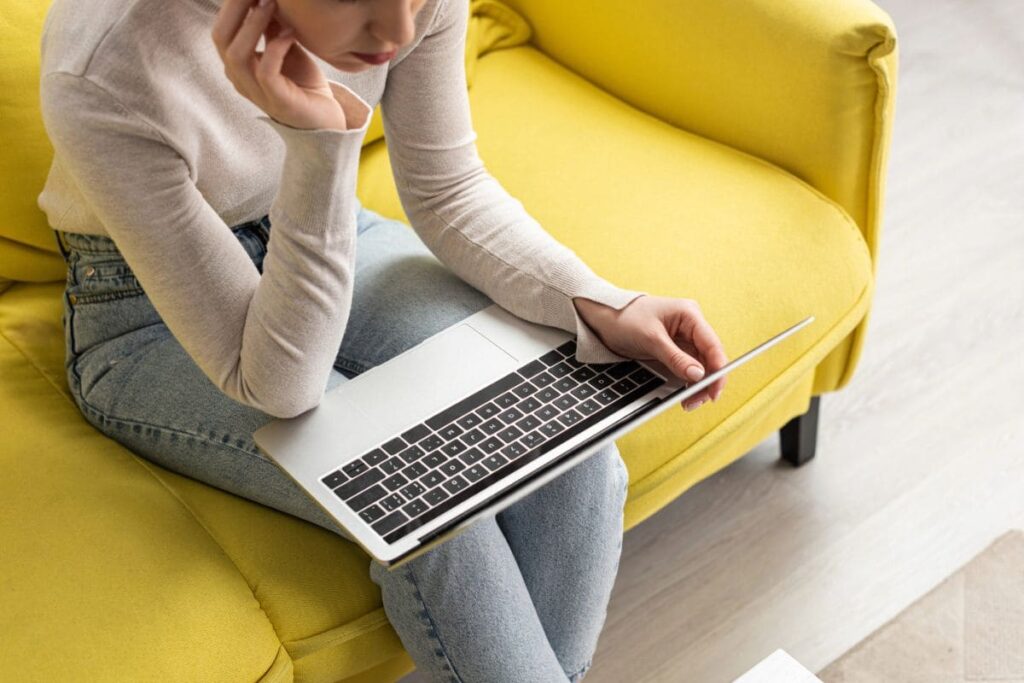 A woman working on a laptop, while sitting on a couch at home.