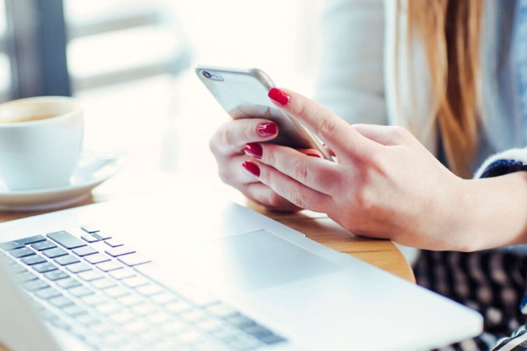 A closeup of a woman's hands holding a cell phone while working from home on a laptop.