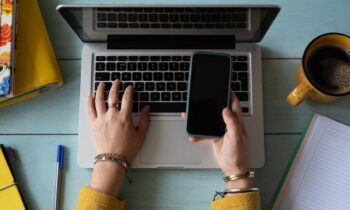 Overhead view of a woman holding a cell phone while working on her laptop.