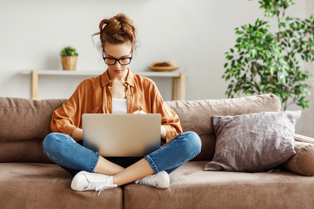 woman wearing glasses sitting cross-legged on sofa working on her laptop to make extra money online